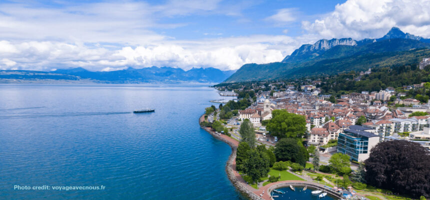 Lakeside town along a blue lake with red-roofed houses, green trees, and mountains in the distance.