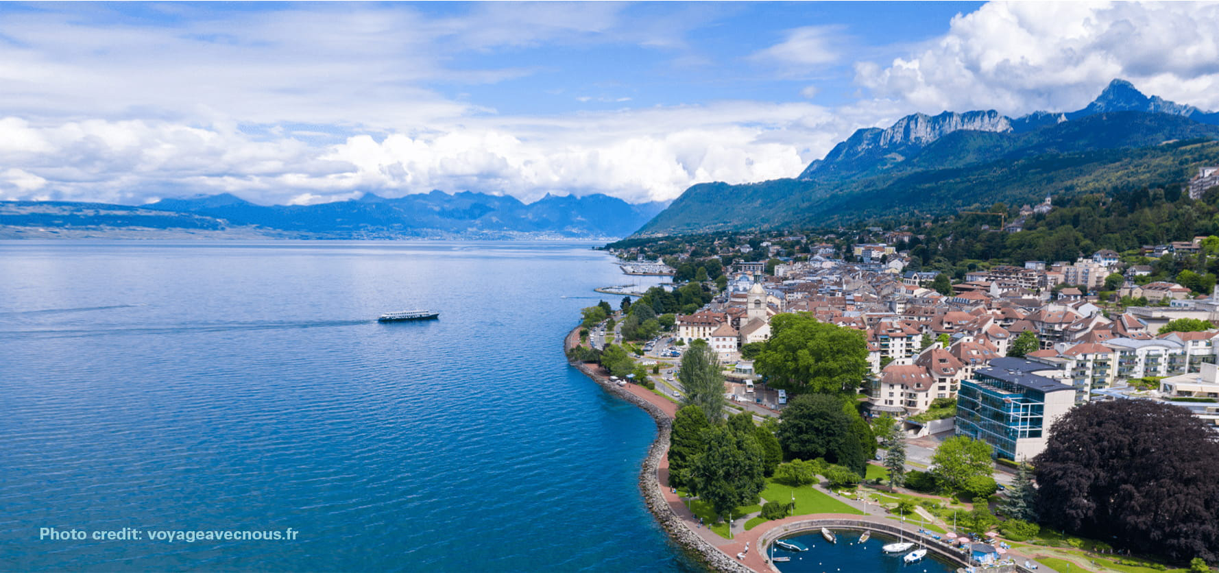 Lakeside town along a blue lake with red-roofed houses, green trees, and mountains in the distance.