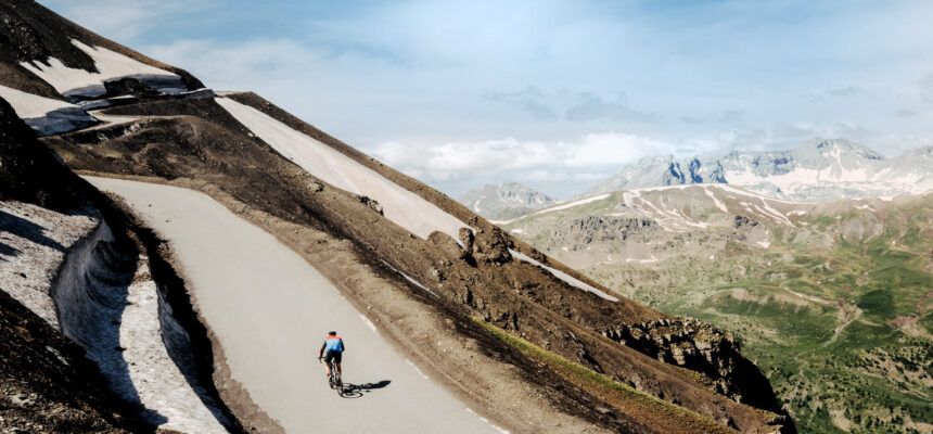Cyclist riding a road bike on a winding mountain pass with rocky slopes and patches of snow under a blue sky.