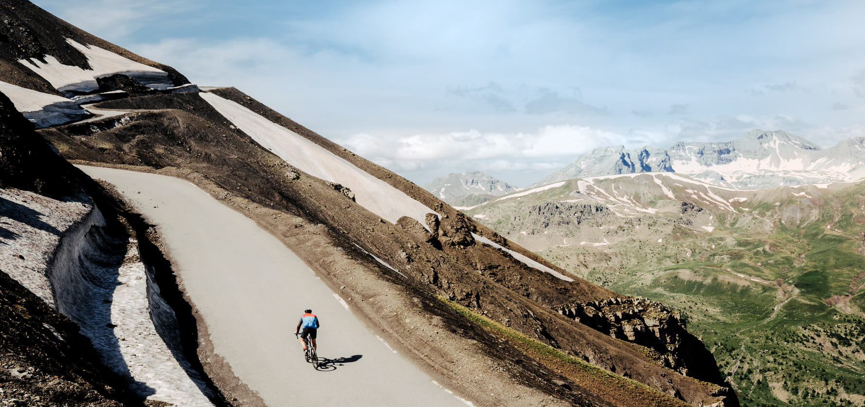 Cyclist riding a road bike on a winding mountain pass with rocky slopes and patches of snow under a blue sky.