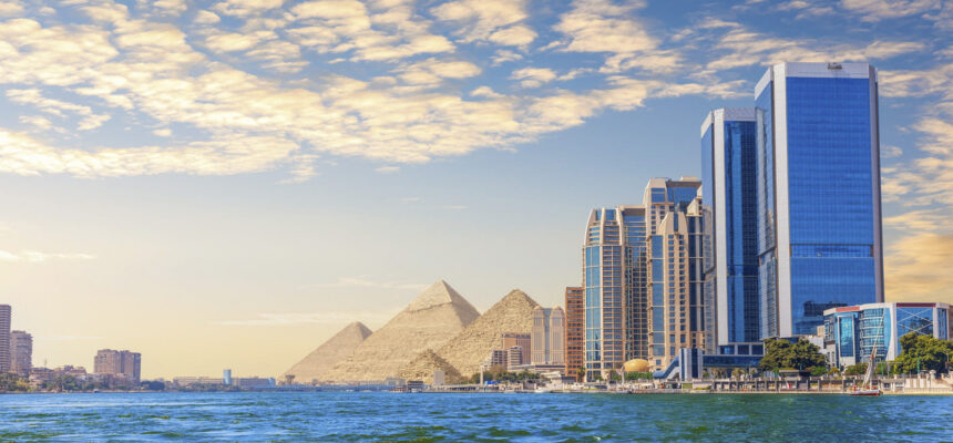 Blue glass skyscrapers along the river with the pyramids in the distance at sunset.